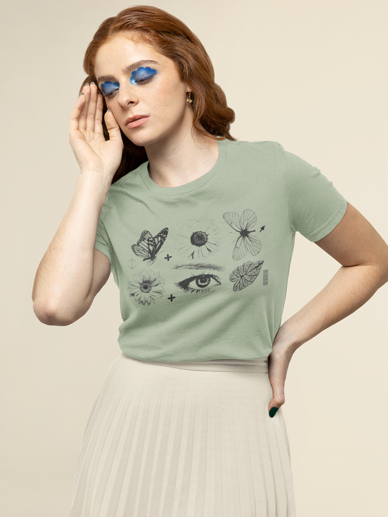 A woman wearing a sage green graphic t-shirt with black line drawings of a butterfly, dandelion, dragonfly, and eye, paired with a pleated cream skirt, against a solid beige background.